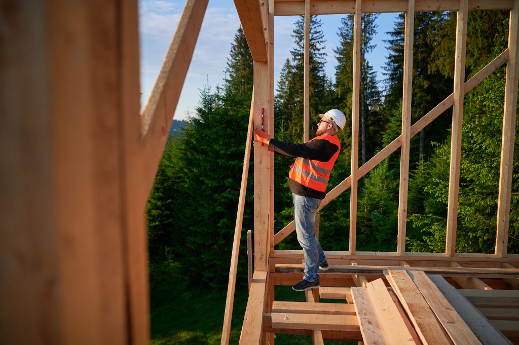 Stavebný špecialista v oranžovej reflexnej veste a bielej prilbe pomocou vodováhy kontroluje rovinu steny montovaného drevodomu zasadeného v zmiešanom lese.