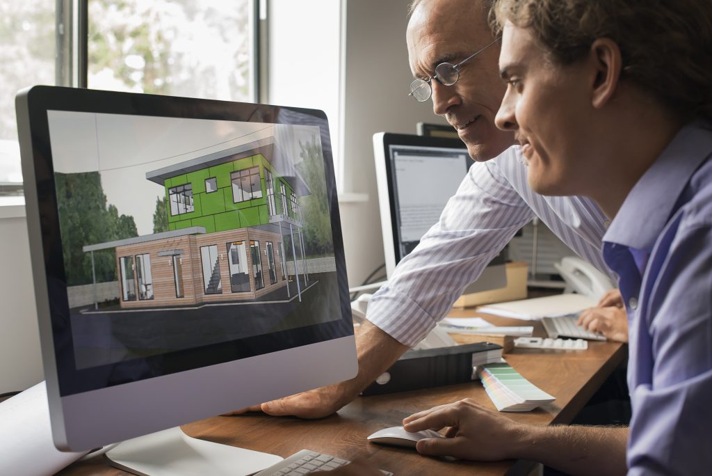 Dvaja architekti pri pracovnom stole pozerajú na monitor iMac s vizualizáciou modernéh dvojposchodového montovaného drevodomu