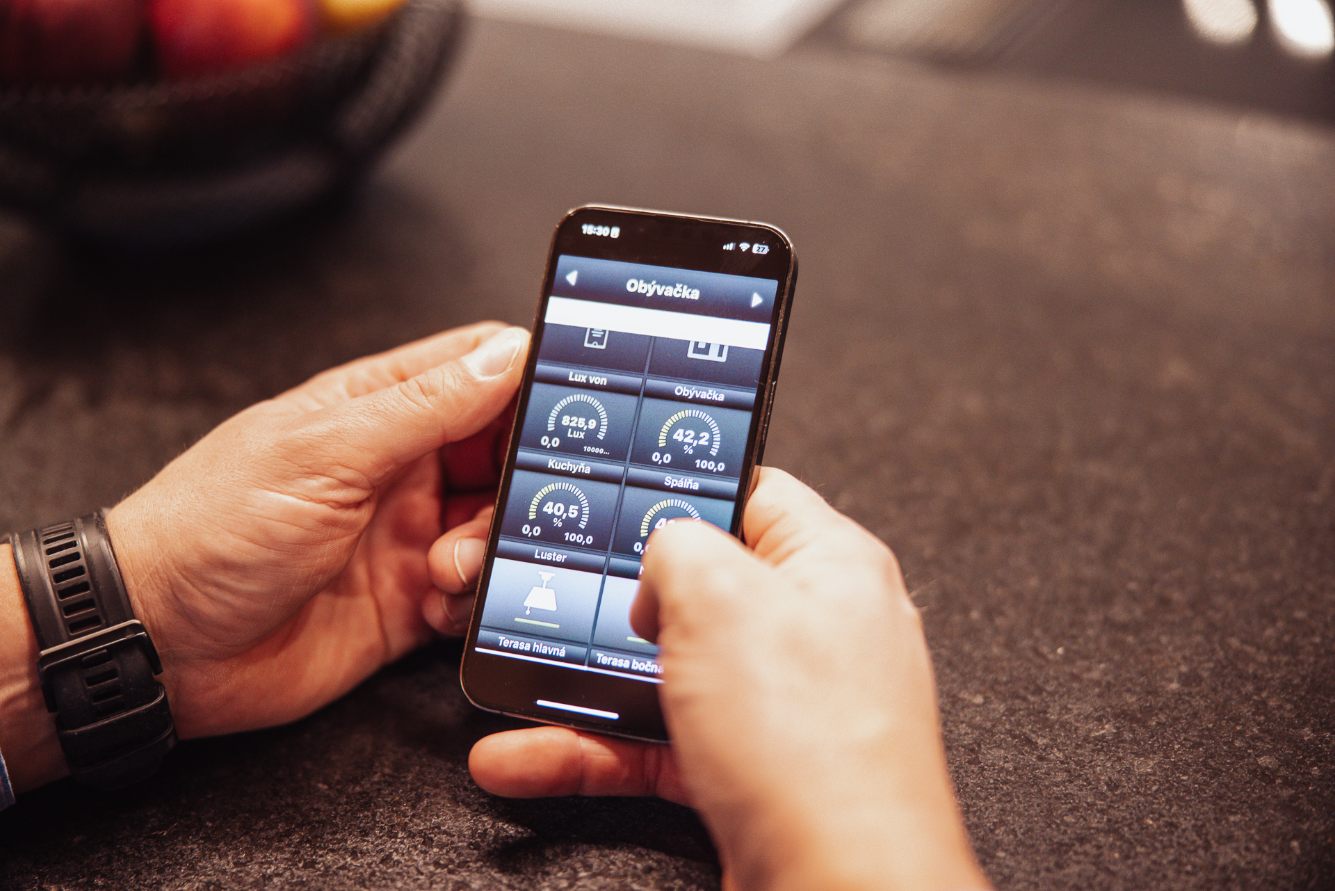 Mužské ruky držia iphone s aktívnou aplikáciou na ovládanie intelignetnej domácnosti s aktívnou obrazovkou s nastavením teploty a ukazovateľmi vlhkosti, svetelnosti a tlaku v miestnosti
